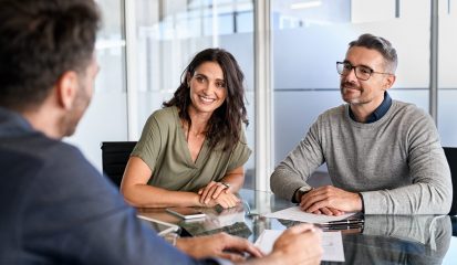 Smiling mature couple meeting with bank manager for investment. Beautiful mid adult woman with husband listening to businessman during meeting in conference room in modern office. Happy middle aged couple meeting loan advisor to buy a new home.